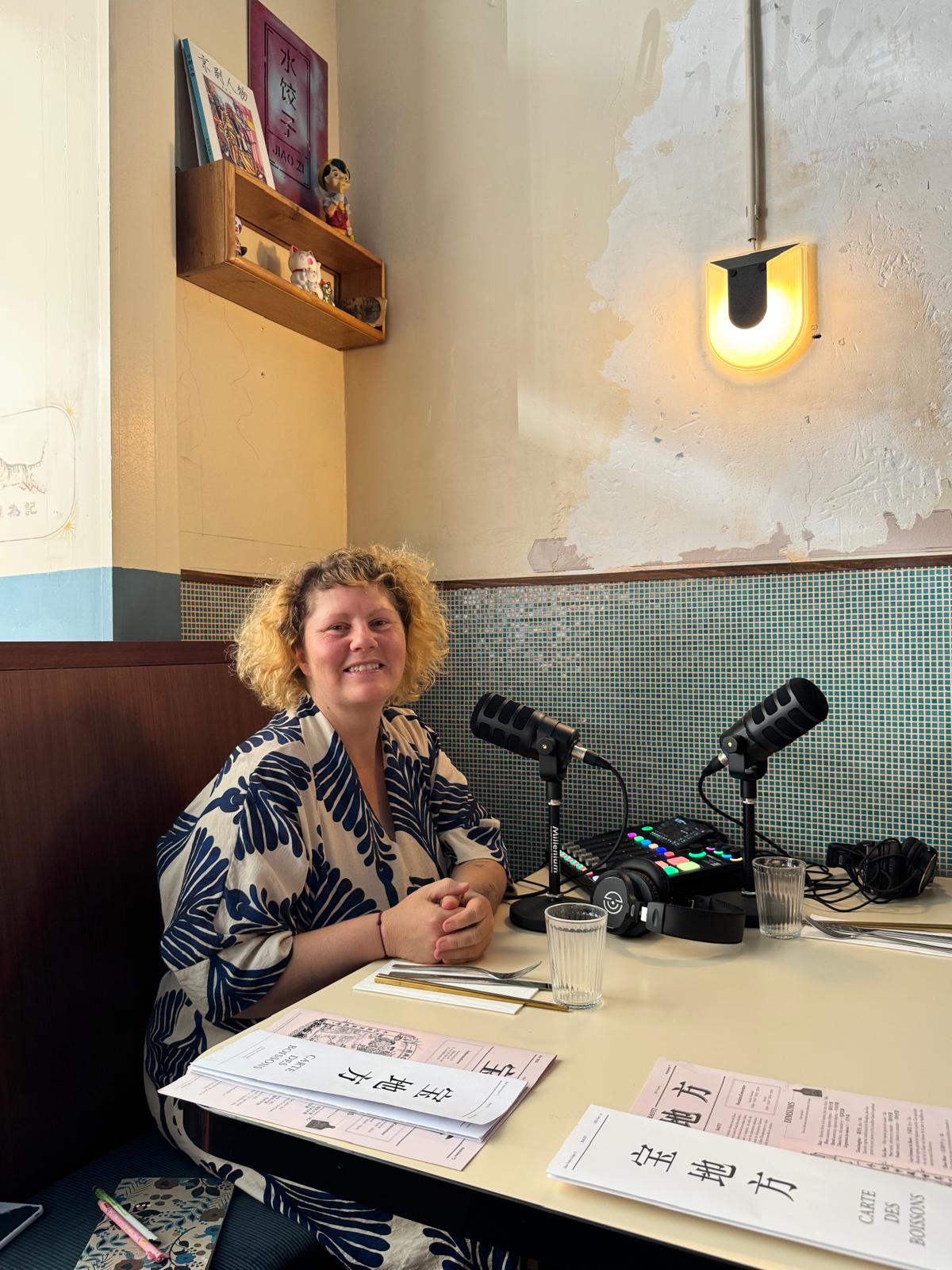 Studio de podcast mobile installé sur une table de restaurant : deux micros, une table de mixage et une femme souriante assise sur la banquette